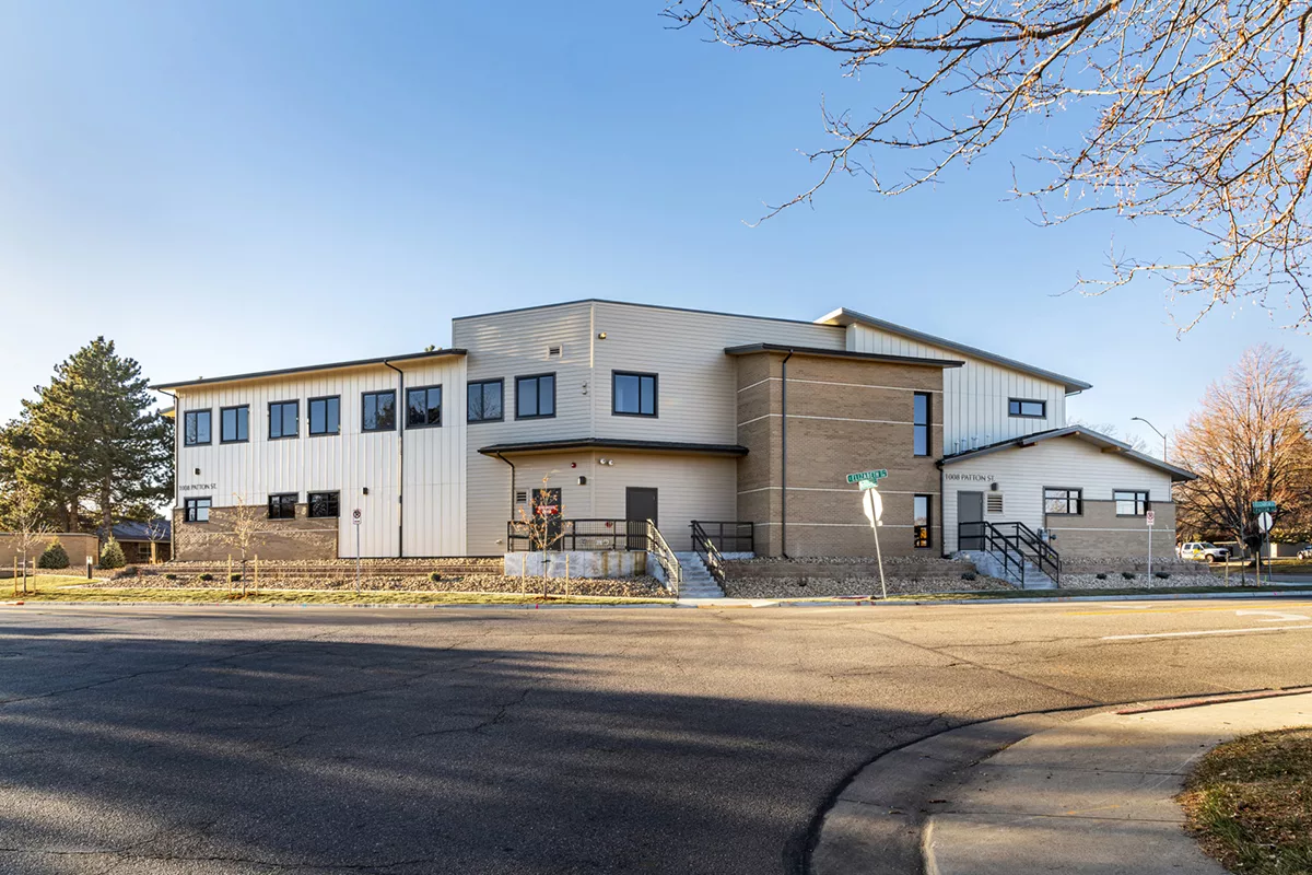 Modern two-story building with beige and white siding, flat roofs, and large windows, located on a street corner with visible sidewalks, steps, and some bare trees around it under a clear blue sky.
