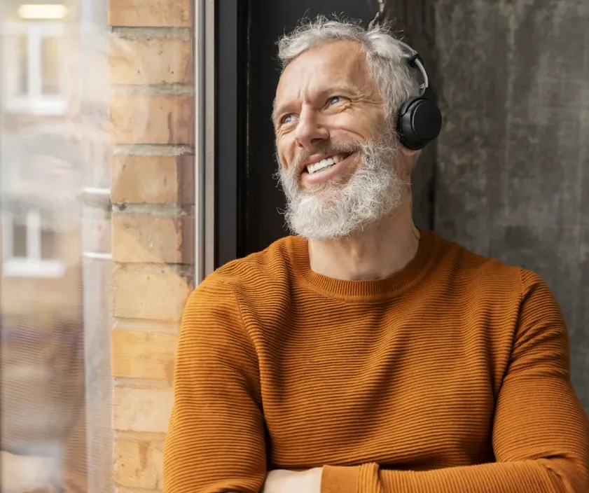 A man with a gray beard wearing a brown sweater and headphones stands by a window, smiling and looking upwards. A brick wall and a building can be seen through the window in the background.