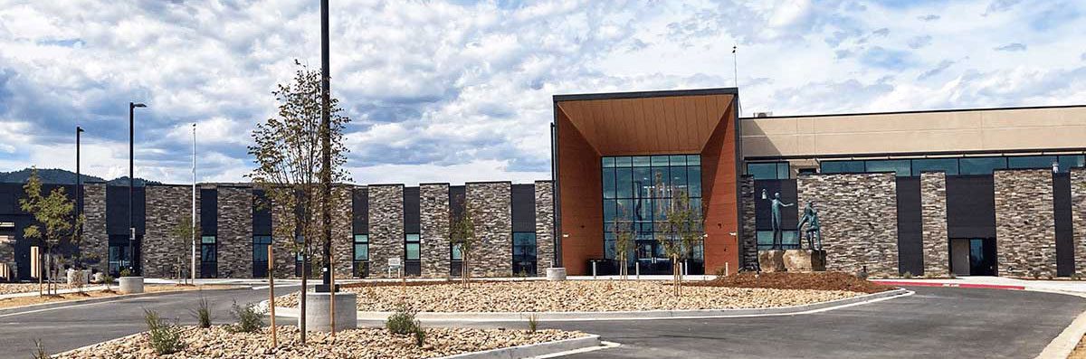 Acute-Care-Facility-Longview-resized Modern building with large windows and stone facade, surrounded by a circular driveway, young trees, and landscaped rocks under a partly cloudy sky.
