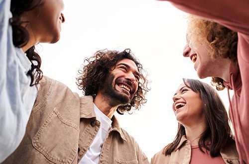 Four people stand close together outdoors, smiling and laughing as they look at each other, creating a joyful and friendly atmosphere. The photo is taken from a low angle against a bright sky.