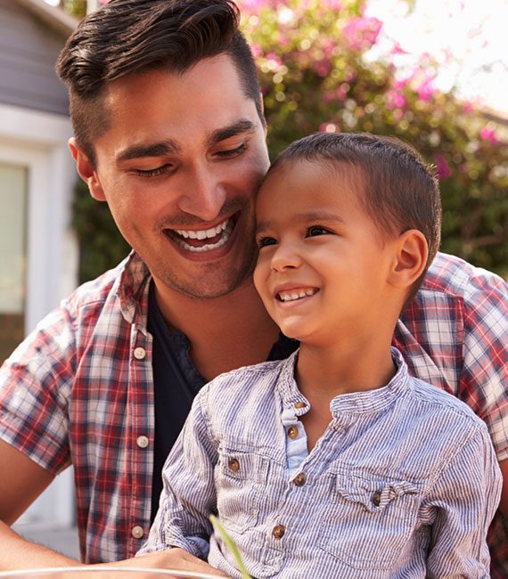 A smiling man holds a young boy on his lap outdoors. Both are laughing and looking to the side, enjoying a sunny day with greenery and pink flowers in the background.
