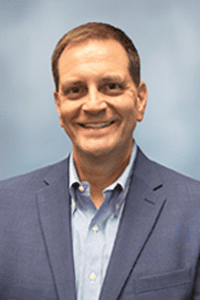 Daniel Peck_COO A man with short brown hair wearing a blue blazer and light blue dress shirt, smiling in front of a plain light blue background.