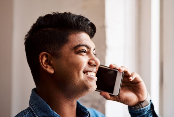 A young man smiling while talking on a smartphone, looking out of a bright window. He is wearing a denim shirt and appears happy and engaged in the conversation.