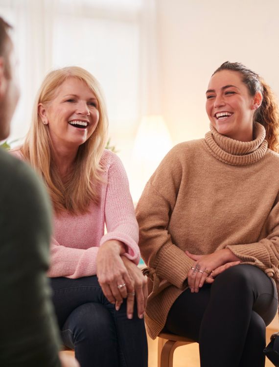 Two women sitting and smiling during a group conversation, both dressed in sweaters. They appear happy and engaged, with blurred figures of other people visible in the foreground. Warm indoor setting.