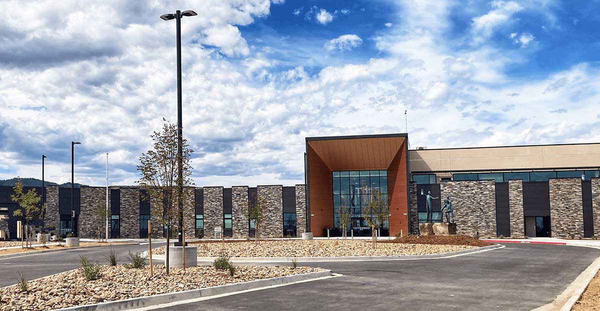 A modern building with stone and glass exterior sits under a partly cloudy sky. The entrance features large windows and angular design. The parking lot and landscaped areas with young trees are in the foreground.