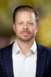 A man with short, light brown hair and a trimmed beard, wearing a dark plaid suit jacket over a white collared shirt, poses in front of a blurred, green and yellow outdoor background.
