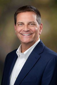 A man with short brown hair wearing a navy blue suit jacket and white dress shirt smiles at the camera against a softly blurred, natural-toned background.