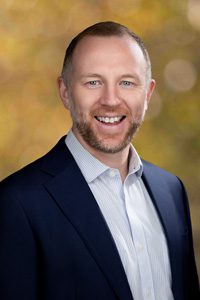 A smiling man with short hair and a beard, wearing a dark suit jacket and light dress shirt, stands in front of a blurred, warm-toned background.