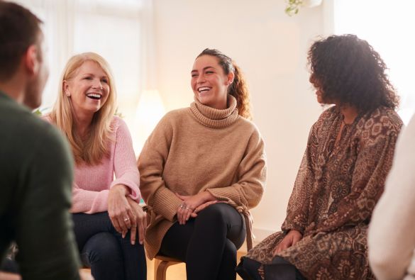 people-in-meeting Four people sit in a circle, smiling and laughing together in a bright, cozy room. The group appears relaxed and engaged in friendly conversation.