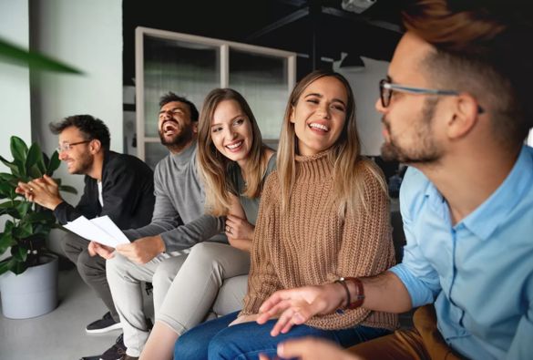 Four people sit together on a sofa, smiling and laughing as they talk. One person holds papers, and a potted plant is visible nearby, creating a friendly, casual atmosphere.