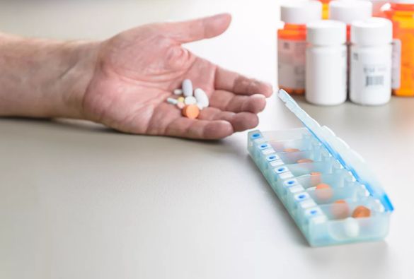 A hand holding various pills above a table with a weekly pill organizer and several prescription medicine bottles in the background.