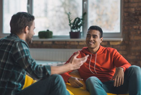 Two men are sitting indoors, engaged in a friendly conversation. One man is wearing a plaid shirt and gesturing with his hand, while the other, in a red hoodie, is smiling and listening. A window and plants are visible in the background.
