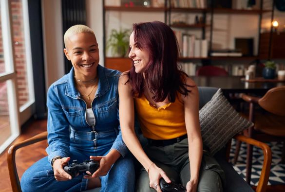 Two women sitting on a couch at home, smiling and holding video game controllers, appearing to enjoy playing a game together in a cozy living room.
