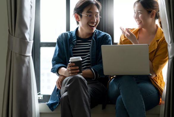 Two people sit by a window, smiling and talking. One holds a coffee cup while the other gestures with one hand and has a laptop on their lap. Light comes in through the window behind them.