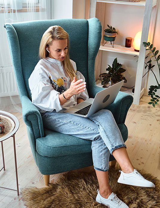 A woman sits in a teal armchair, gesturing with her hand while looking at a laptop on her lap. She wears jeans, a white blouse, and white sneakers. There are plants and shelves in the background.