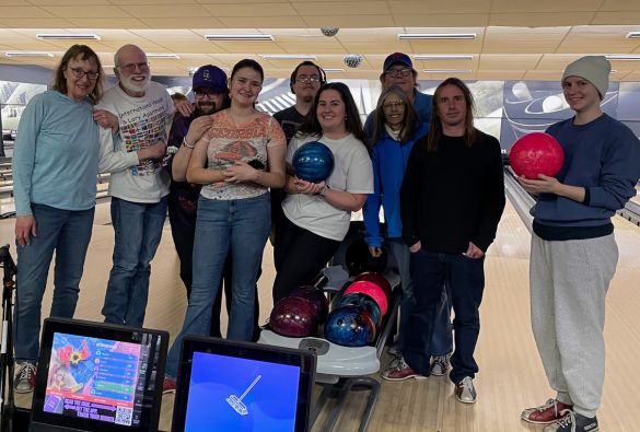 A group of nine people stand together at a bowling alley, some holding bowling balls, smiling at the camera. Bowling lanes, pins, and ball return machines are visible in the background.