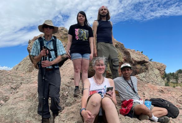 Five people pose on rocky terrain under a blue sky with scattered clouds. Two are seated in front, three stand behind them. They wear casual outdoor clothing, some holding water bottles and hiking gear.
