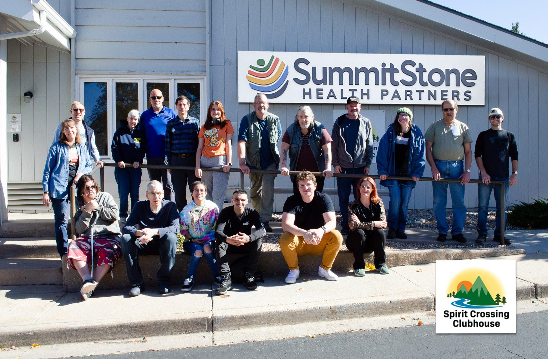 A group of people pose together outside the SummitStone Health Partners building, standing and sitting near a handrail, next to a sign that reads "Spirit Crossing Clubhouse.