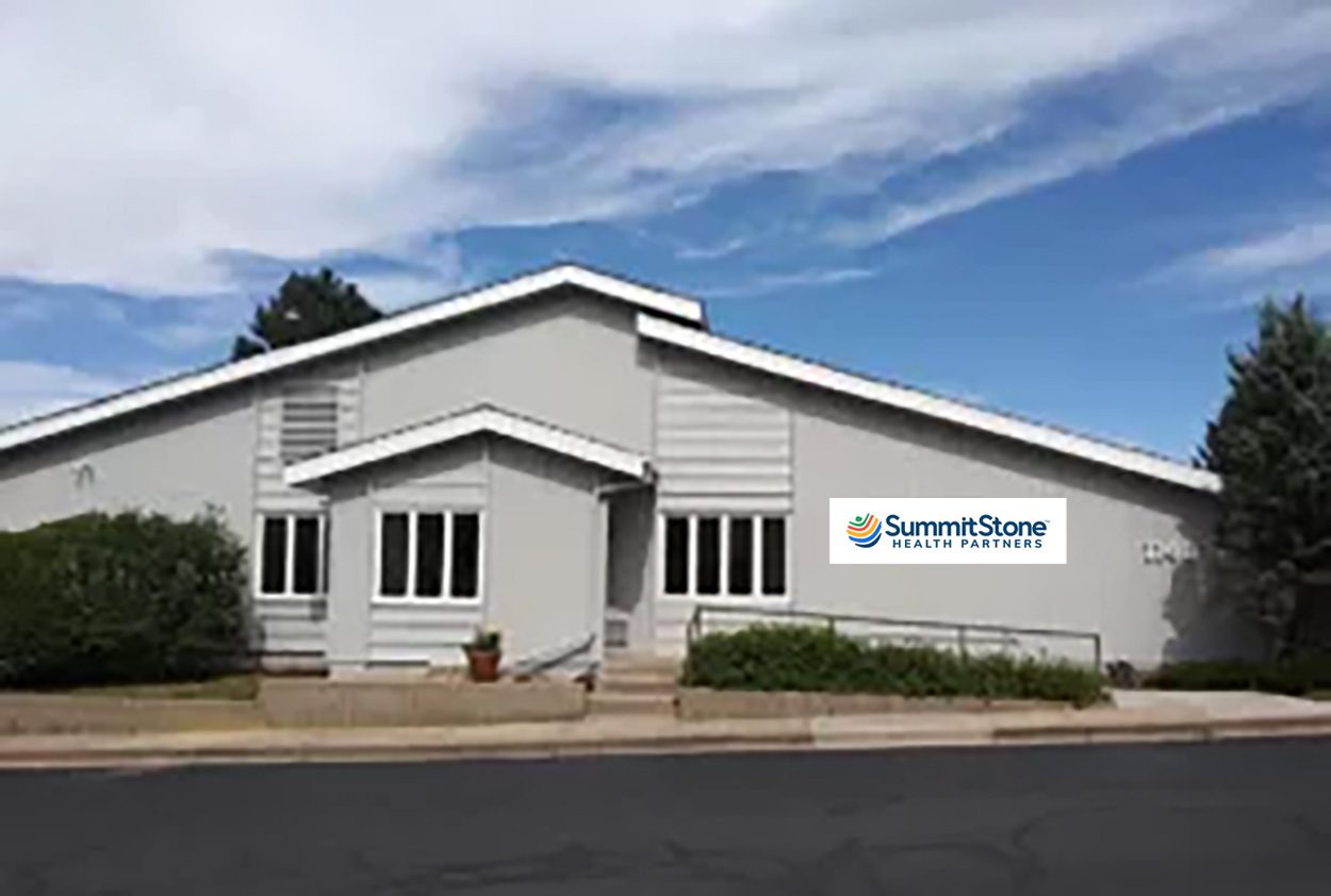 A single-story gray building with white trim, a ramp and steps leading to the entrance, and a "SummitStone Health Partners" sign on the front. Trees and a cloudy blue sky are in the background.