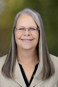 A middle-aged woman with long straight gray hair and glasses is smiling. She is wearing a beige blazer over a black top, with a blurred green background.