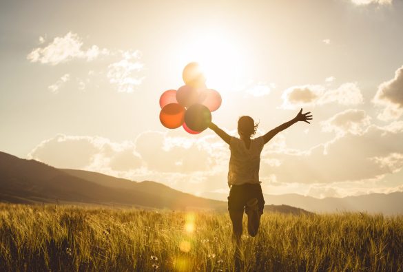 A person runs through a grassy field at sunset, holding colorful balloons with arms outstretched. The sun shines brightly in the sky, creating a warm and joyful atmosphere.